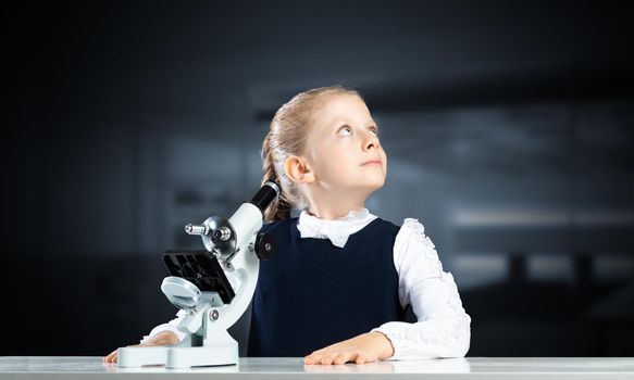 Little girl scientist with microscope on blurred background. Research and discovery concept with copy space. Elementary science class in modern school. Schoolgirl in schoolwear sitting at desk.