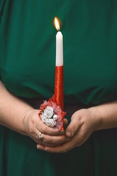 The hands of a woman in a green dress hold a large burning decorative candle, close-up.