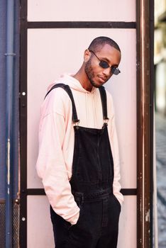 Young black man wearing casual clothes and sunglasses, against a pink urban background. African guy with bib pants standing outdoors