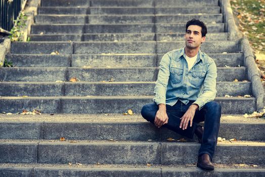 Portrait of a young handsome man, model of fashion, with formal hairstyle sitting in urban stairs, wearing casual clothes. Blue eyes guy in urban background.