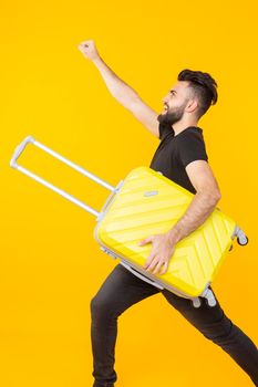 Cute pretty young man with a beard holding a yellow suitcase in his hands on a yellow background. Concept of travel and vacations. Advertising space