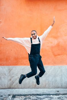 Young black man wearing casual clothes jumping in urban background. Lifestyle concept. Millennial african guy with bib pants outdoors