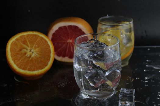 splashes of water over a glass of water and ice. Nearby are fruits orange and grapefruit, ice. On a black background. Refreshing cold ice drink.