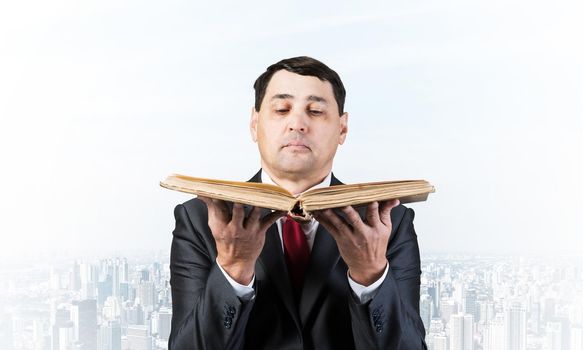 Serious businessman holding open old book. Adult man in business suit standing on cloudy cityscape background. Lawyer reading big legal regulation book. Professional business accounting and consulting