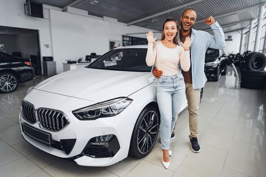 Happy couple of caucasian woman and african american man standing near their new luxury car inside car salon