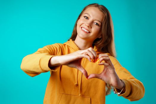 Smiling young woman showing heart sign with her hands against blue background