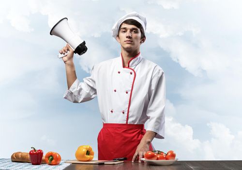 Young chef standing with megaphone in hand. Emotional caucasian chef in white hat and red apron on blue sky background. Restaurant advertisement, food retail campaign announcement. Catering service