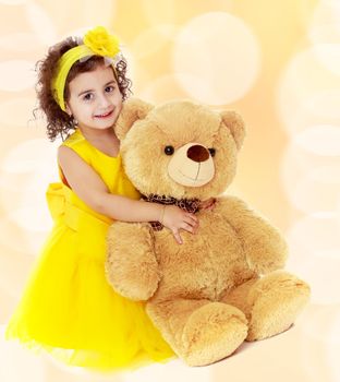 Joyful little girl in a yellow dress and bow on her head sitting on the floor. Girl hugging a big Teddy bear.Winter brown abstract background with white snowflakes.