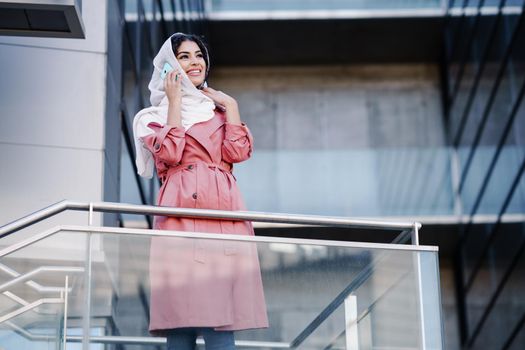 Young Arab woman walking on the street wearing hijab headscarf using smart phone in a office building. Business background.