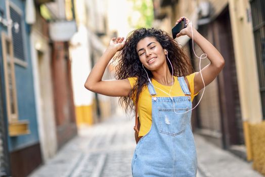 Attractive African girl listening to music with earphones outdoors. Arab woman in casual clothes with curly hairstyle in urban background with closed eyes.