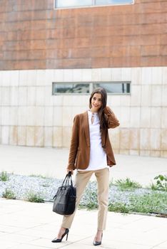 Young businesswoman standing outside of office building with briefcase. Beautiful woman wearing formal wear. Young girl with brown jacket and trousers.
