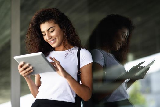 Young North African woman with looking at her digital tablet outdoors. Arab girl using a device reflected in window glass.