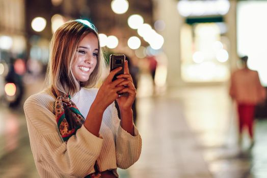 Blonde woman taking photograph with smartphone at night in the street. Defocused city lights at the background. Pretty girl with pigtail hairstyle at night smiling.