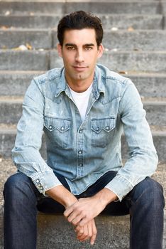 Portrait of a young handsome man, model of fashion, with formal hairstyle sitting in urban stairs, wearing casual clothes. Blue eyes guy in urban background.