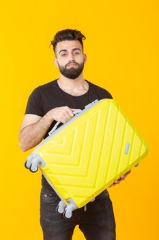 Cute pretty young man with a beard holding a yellow suitcase in his hands on a yellow background. Concept of travel and vacations. Advertising space