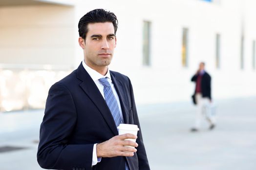 Businessman with formal clothes drinking coffee to go with a take away cup. Man wearing blue suit and tie in urban background.
