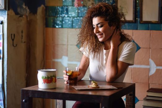Young arabic smiling woman with black curly hairstyle sitting in a beautiful bar with vintage decoration. Arab girl in casual clothes drinking a soda.