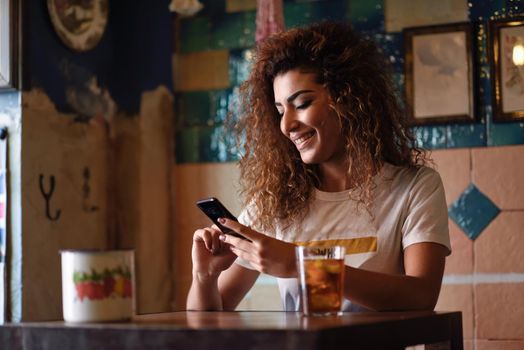 Young arabic woman with black curly hairstyle sitting in a beautiful bar with vintage decoration. Arab girl in casual clothes drinking a soda and looking at her smart phone