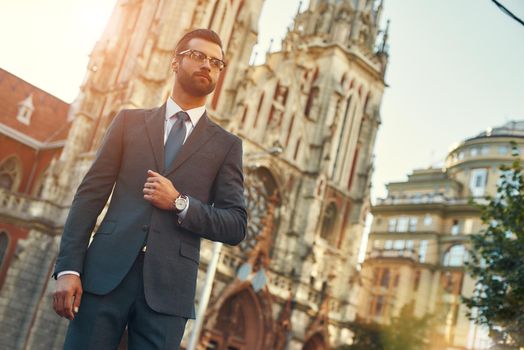 Great day Confident and stylish businessman in full suit and eyeglasses looking away while standing against beautiful church. Business concept. Stylish people