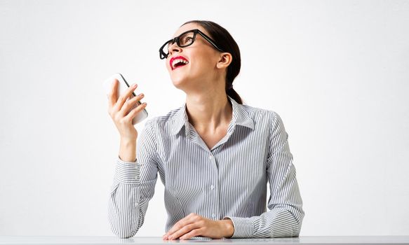 Smiling young woman sits at desk and dreamy looking upward. Portrait of beautiful girl in glasses with bright red lips on white wall background. Human resource and career development concept.