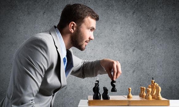 Businessman moving chess figure in chessboard. Successful management and leadership. Handsome man in business suit at desk with chess on grey wall background. Operative tactics and strategy planning.