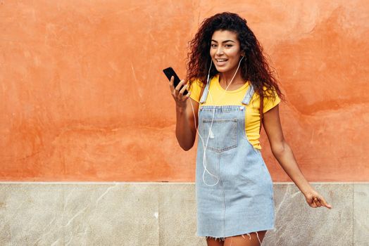 Happy North African woman listening to music with earphones outdoors. Arab girl in casual clothes with curly hairstyle on urban wall.