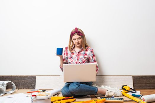 Beautiful girl using laptop on floor at home. Apartment remodeling and house interior renovation concept with copy space. Young attractive woman in red checkered shirt and jeans relaxing with coffee.