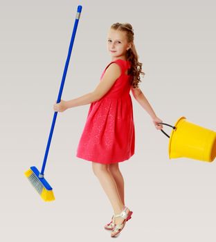 Beautiful little girl in a red short dress with a bucket and brush pleasure to cleans the house.On a gray background.