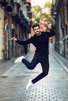 Young bearded man jumping in urban street wearing casual clothes. Happy guy with beard and modern hairstyle.