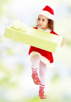 Cute little curly girl in a coat and hat of Santa Claus carries the big green box. A girl holds a box with his knee.white-green blurred abstract background with snowflakes.