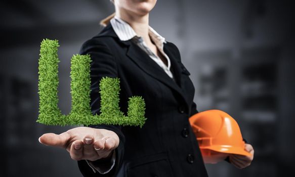 Businesswoman holds in palm green plant in shape of grow up chart. Woman engineer in business suit with orange safety helmet. Engineering and project management. Money saving and investing.