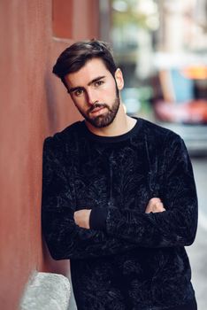 Young bearded man, model of fashion, looking at camera in urban background wearing casual clothes. Guy with beard and modern hairstyle in the street.