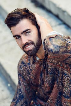 Young smiling man, model of fashion, wearing modern shirt in the street. Guy with beard and modern hairstyle in urban background.