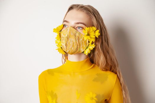 Slim female model in protective mask with fresh yellow flowers standing in transparent outfit on white background in studio