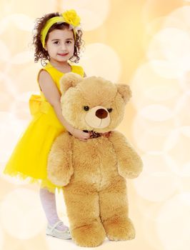 Joyful little girl Princess, dressed in a yellow dress, hugging a big Teddy bear. Standing at full height.Winter brown abstract background with white snowflakes.