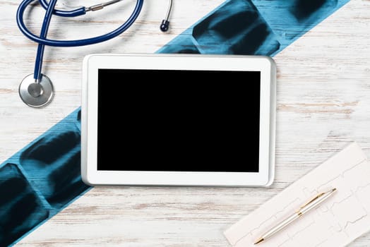 Laboratory result of patient examination lies on doctor table. Top view tablet computer with blank screen, x-ray image, stethoscope and cardiogram on wooden desk. Digital technology in cardiology.