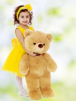 Joyful little girl Princess, dressed in a yellow dress, hugging a big Teddy bear. Standing at full height.white-green blurred abstract background with snowflakes.
