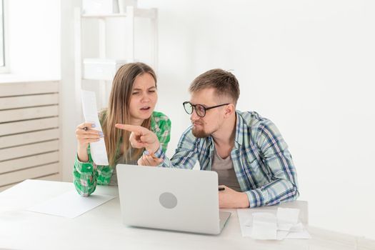 Young husband and wife are upset about numerous checks for payments and purchases for repairs while sitting at the table with a laptop. The concept of low living standards and savings. Copyspace