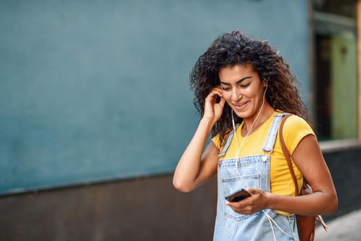 Attractive African woman listening to music with earphones outdoors. Black girl in casual clothes with curly hairstyle in urban background.