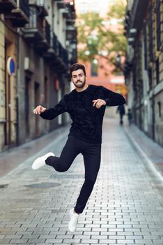 Young bearded man jumping in urban background with open arms wearing casual clothes. Guy with beard and modern hairstyle in the street.
