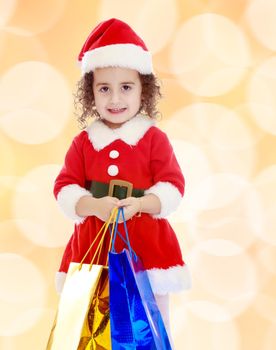 Gentle little curly-haired girl in a coat and hat of Santa Claus holding colorful shopping bags. Close-up.Winter brown abstract background with white snowflakes.