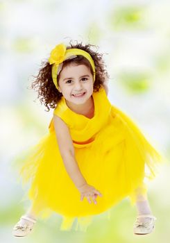 Happy little curly girl in a bright yellow dress, dancing happily.white-green blurred abstract background with snowflakes.