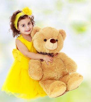 Joyful little girl in a yellow dress and bow on her head sitting on the floor. Girl hugging a big Teddy bear.white-green blurred abstract background with snowflakes.