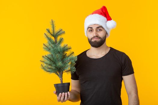 Handsome young man holding a red gift box and christmas tree in his hands posing in a New Year's cap on a yellow background. Merry Christmas and Happy New Year greetings concept