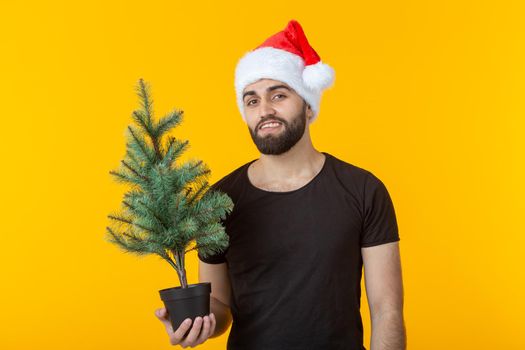 Handsome young man holding a red gift box and christmas tree in his hands posing in a New Year's cap on a yellow background. Merry Christmas and Happy New Year greetings concept