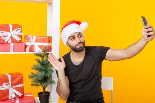 Young handsome hipster man in a santa claus hat making a selfie on a smartphone on the background of greeting gifts and a New Year tree on a yellow background. New Year and Christmas concept.