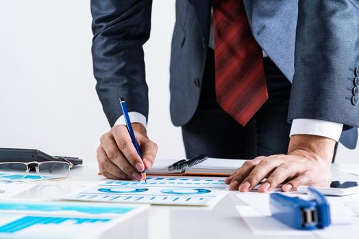 Businessman standing near office desk and analyzing financial charts. Successful corporate manager in business suit and tie. Close-up man hand with pen. Entrepreneurship and leadership concept.