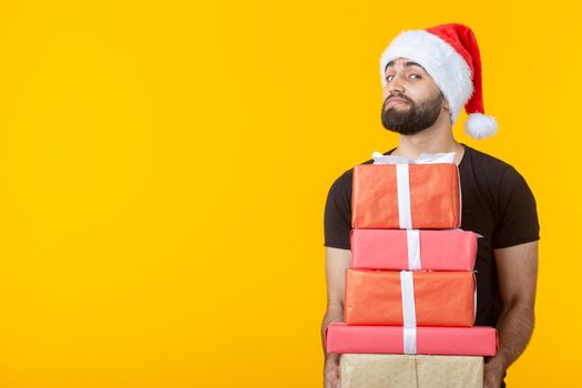 Disgruntled young man with a beard in a Santa Claus hat holds five gift boxes posing on a yellow background. Concept of gifts and greetings for Christmas and New Year