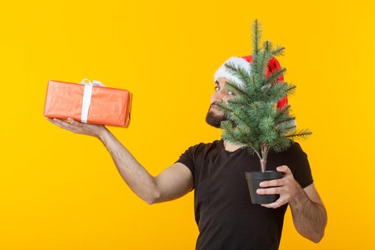 Handsome young man holding a red gift box and christmas tree in his hands posing in a New Year's cap on a yellow background. Merry Christmas and Happy New Year greetings concept