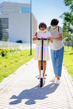 Mother teaching her daughter to ride an electric scooter in urban background wearing surgical masks